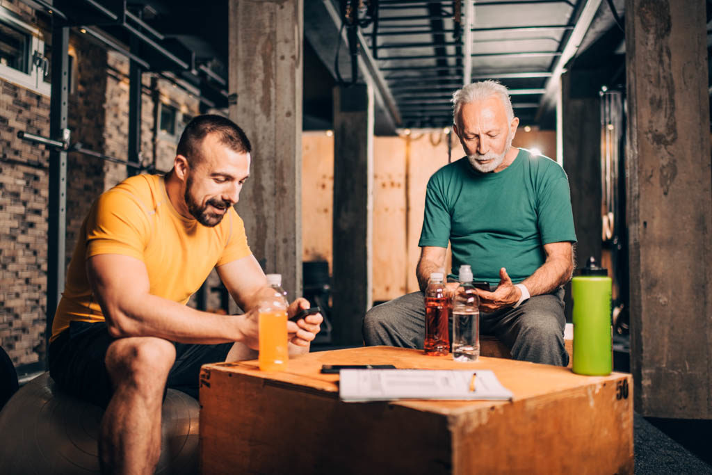 An older client and a younger personal trainer sit and log a workout with coaching software in a gym.
