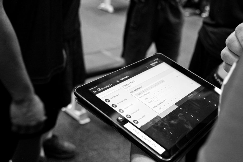 A tablet surrounded by athletes shows a screen from the coaching platform TrainHeroic.