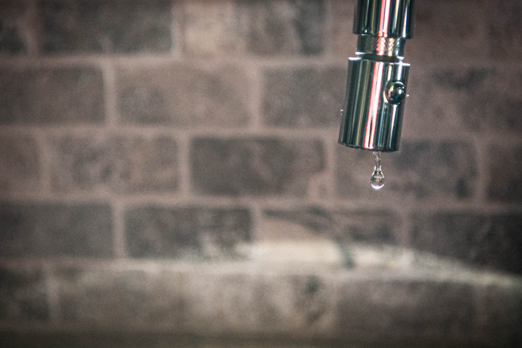 A close-up shot of a droplet of water falling from a kitchen faucet with a brick backsplash.