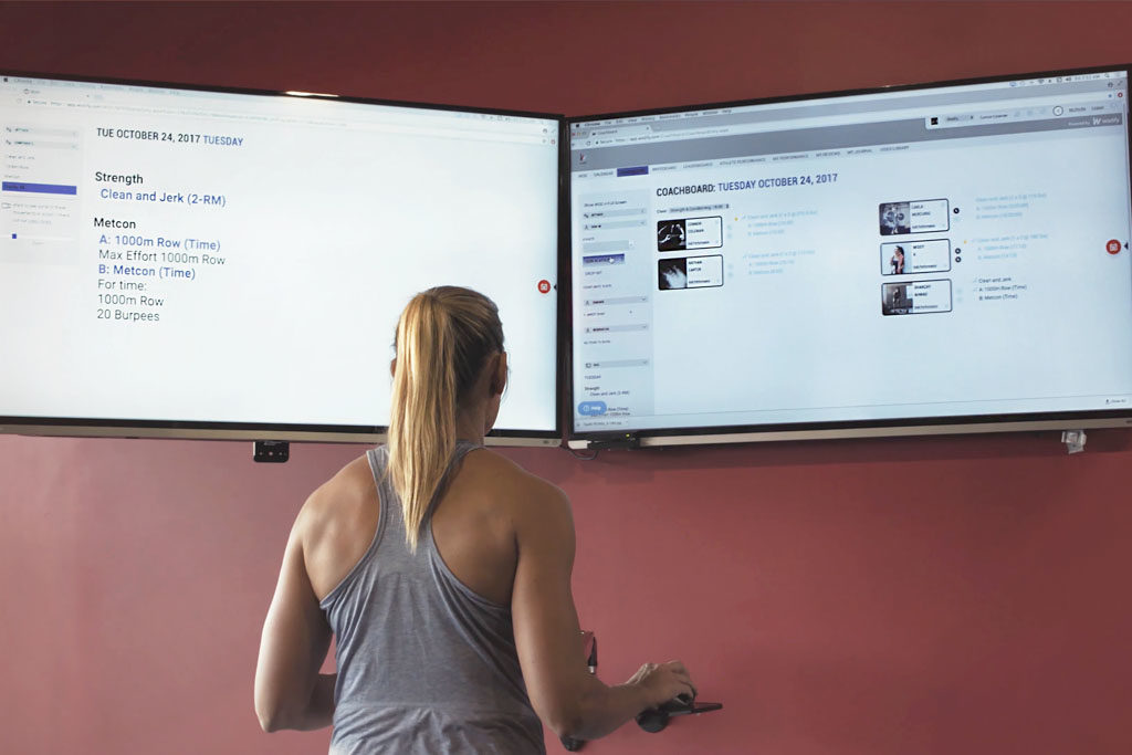 A blond woman in a tank top stands in front of two flat screens displaying the coaching software Wodify.