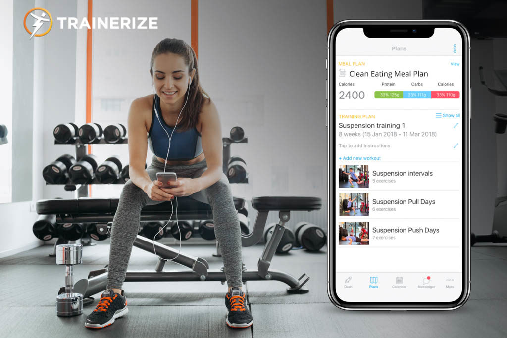 A woman in tights and a sports bra sits on a gym bench and enters workout details using Trainerize on a mobile device.