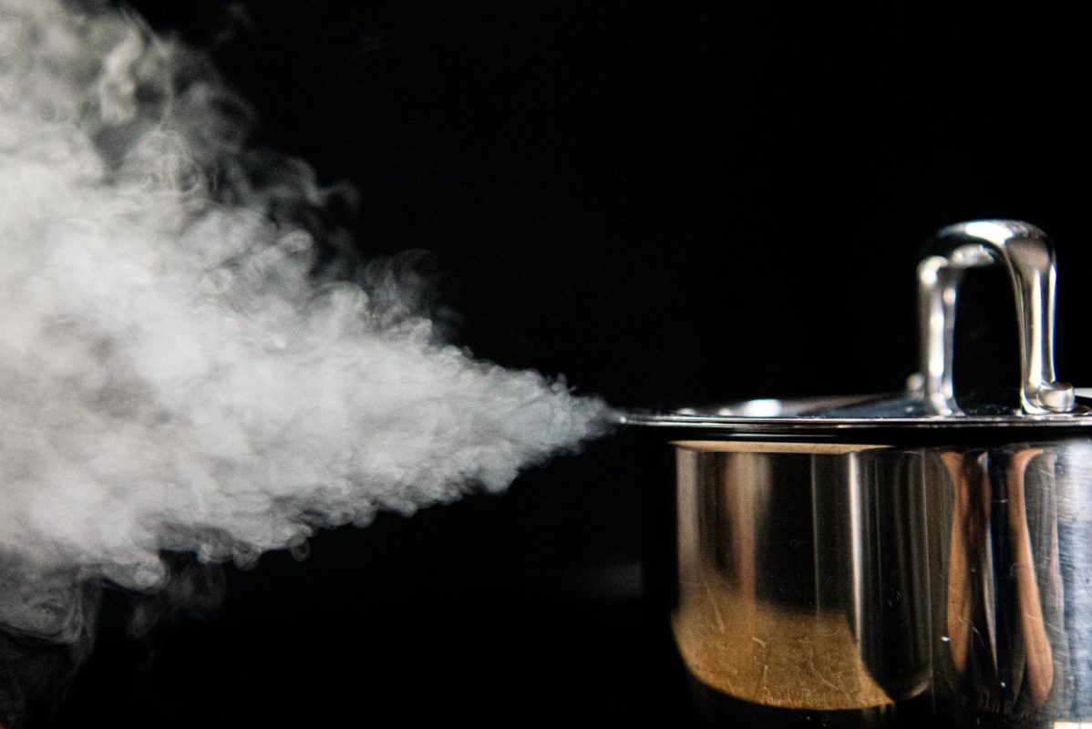 On a black background, a white cloud of steam shoots out the side of a silver pot full of boiling water.