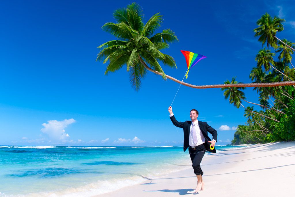 A man in a black suit and a tie runs along a tropical beach while flying a multi-colored kite overhead.