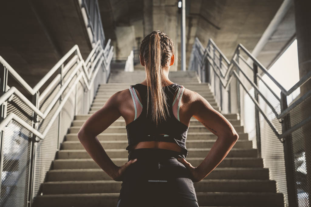 A woman in workout wear stands with her hands on her hips and looks up at a concrete staircase rising in front of her.