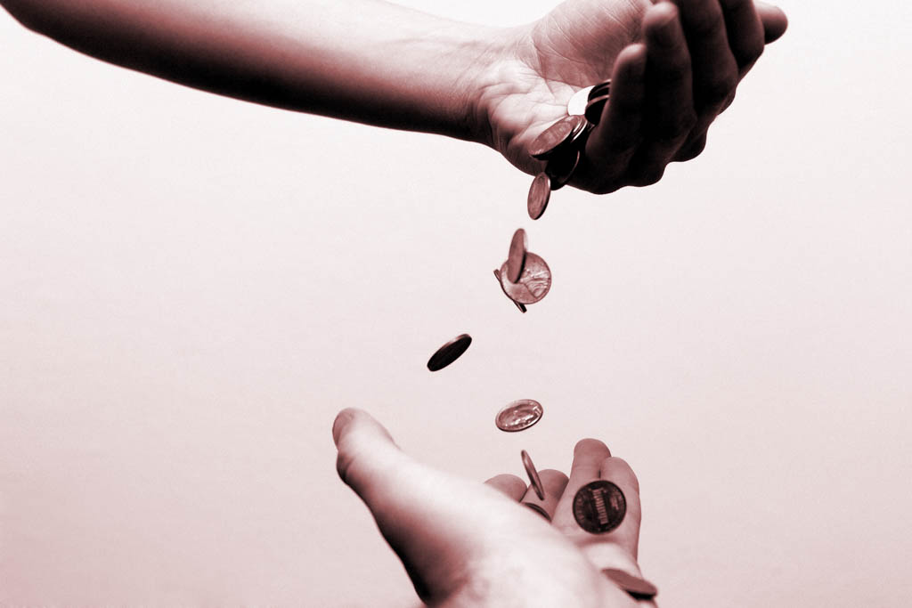 To illustrate the article "What Is Wealth," a hand drops coins into another hand below.