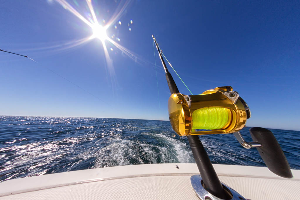 A fishing rod with a gleaming gold reel sits in a holder on a boat with a brilliant blue sky and sun above an ocean with small whitecaps.