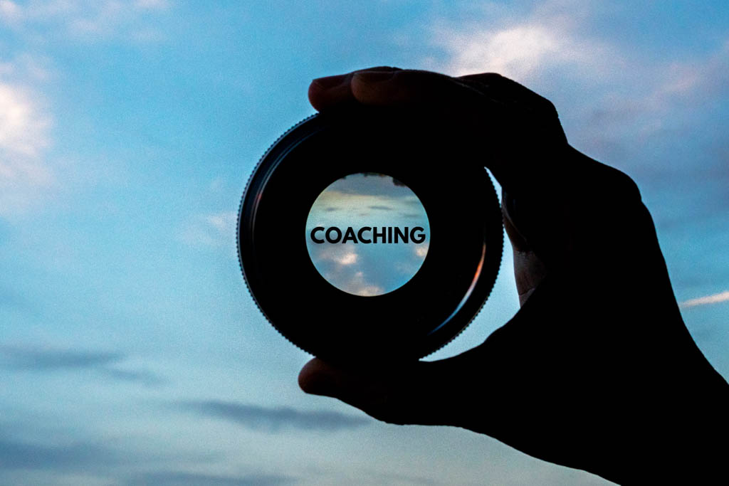 A silhouette of a hand holding a lens against a blue sky, with the word "coaching" at the focal point of the lens.