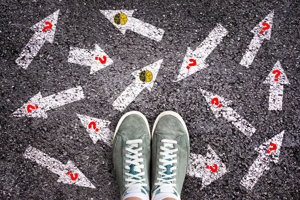 Two feet in running shoes stand on asphalt covered with a series of confusing white arrows with red question marks on them.