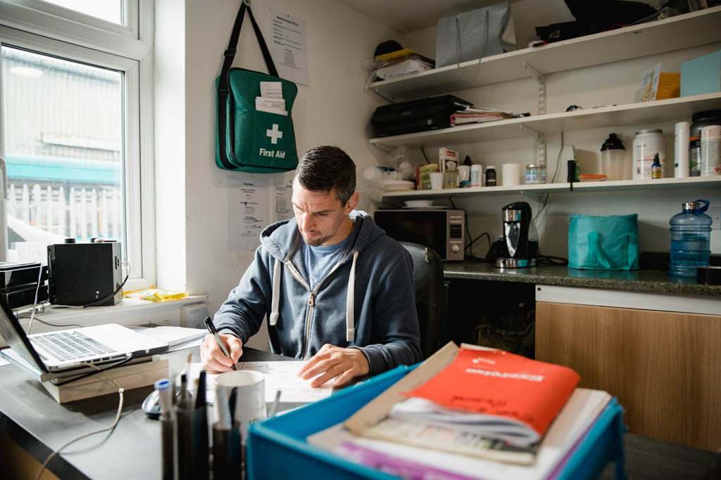 A trainer in a blue hoodie sits in front of a desk in a gym office and studies fitness books.