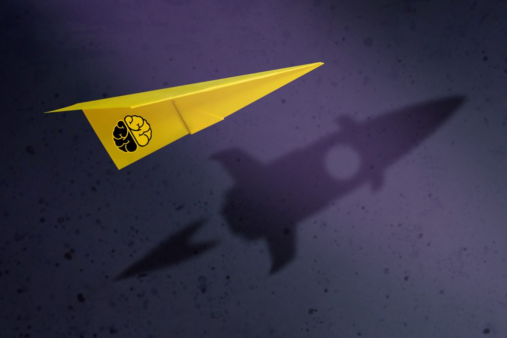A yellow paper airplane with the Two-Brain Business logo casts a shadow that looks like a rocket ship on a purple wall.