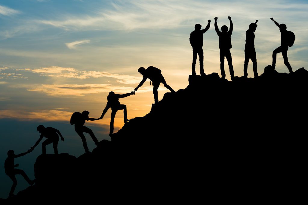 a group of silhouettes help each other up the side up a mountain to stand cheering together at the top against a sunset.