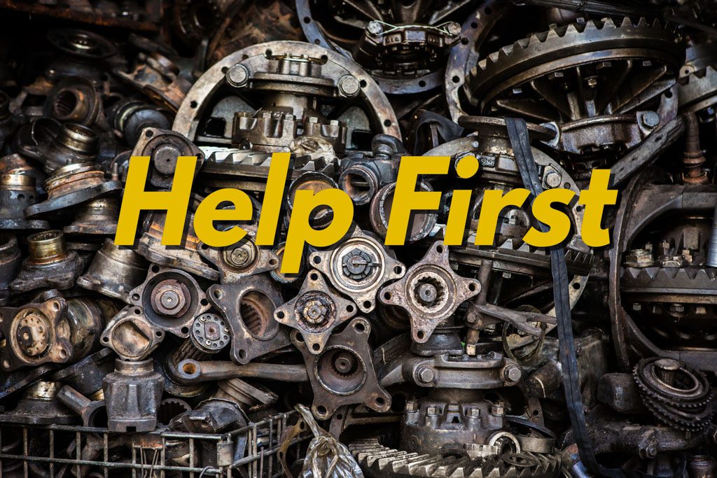 The gold words "help first" are superimposed over a huge pile of old automobile engine parts.