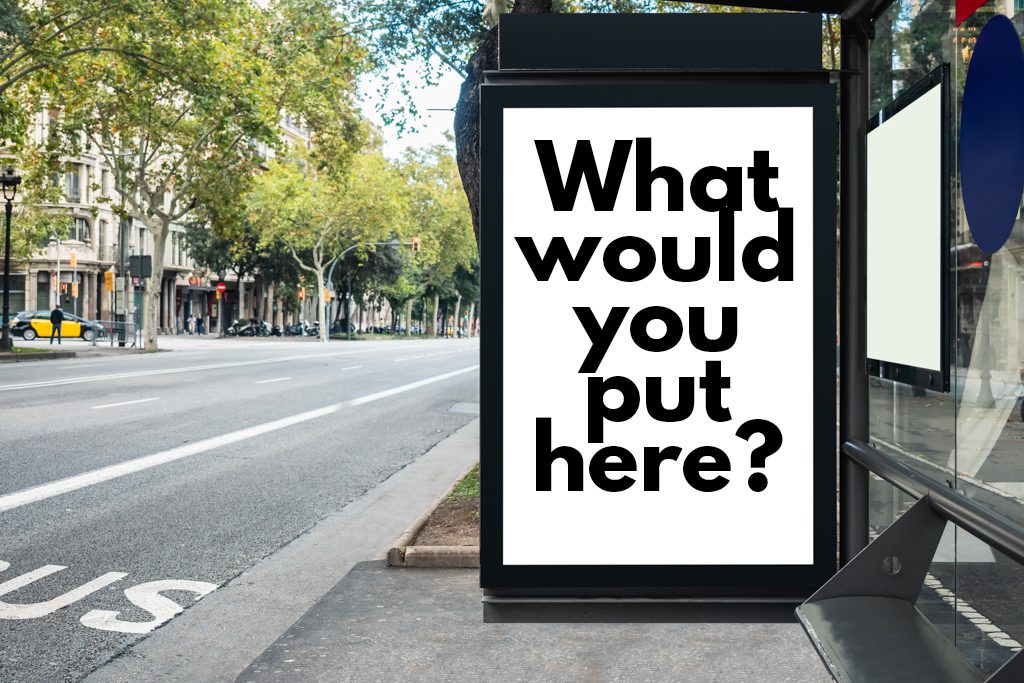 On a deserted, tree-lined street, a bus shack sign that reads "what would you put here?"