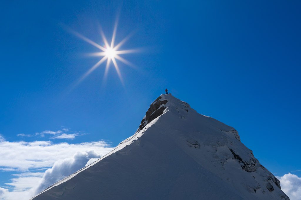 A lone figure stands atop a great snowy mountain against a bright blue sky with a sunburst and a few white clouds.