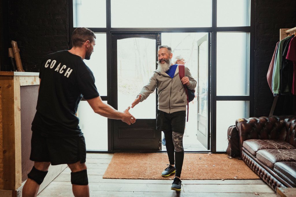 A credentialed personal trainer meets a new client at the door to the gym with a handshake.