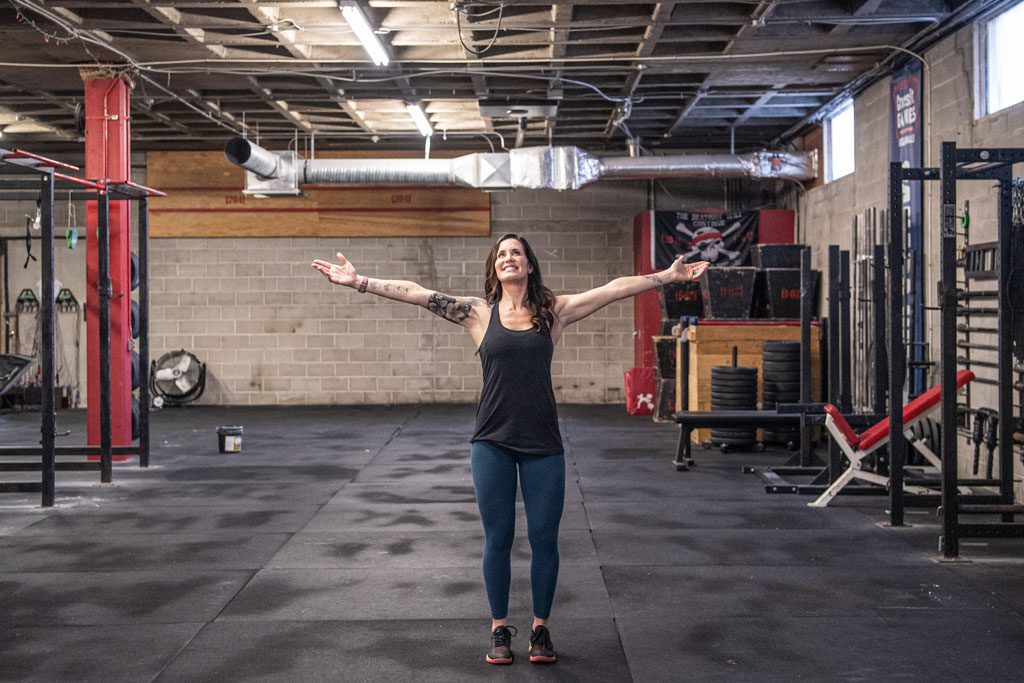 A gym owner with long brown hair stretches her arms out and looks up to smile in her gym.