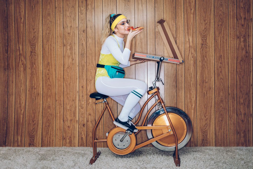 A woman in bright 1980s fitness attire eats a pizza while riding an exercise bike.
