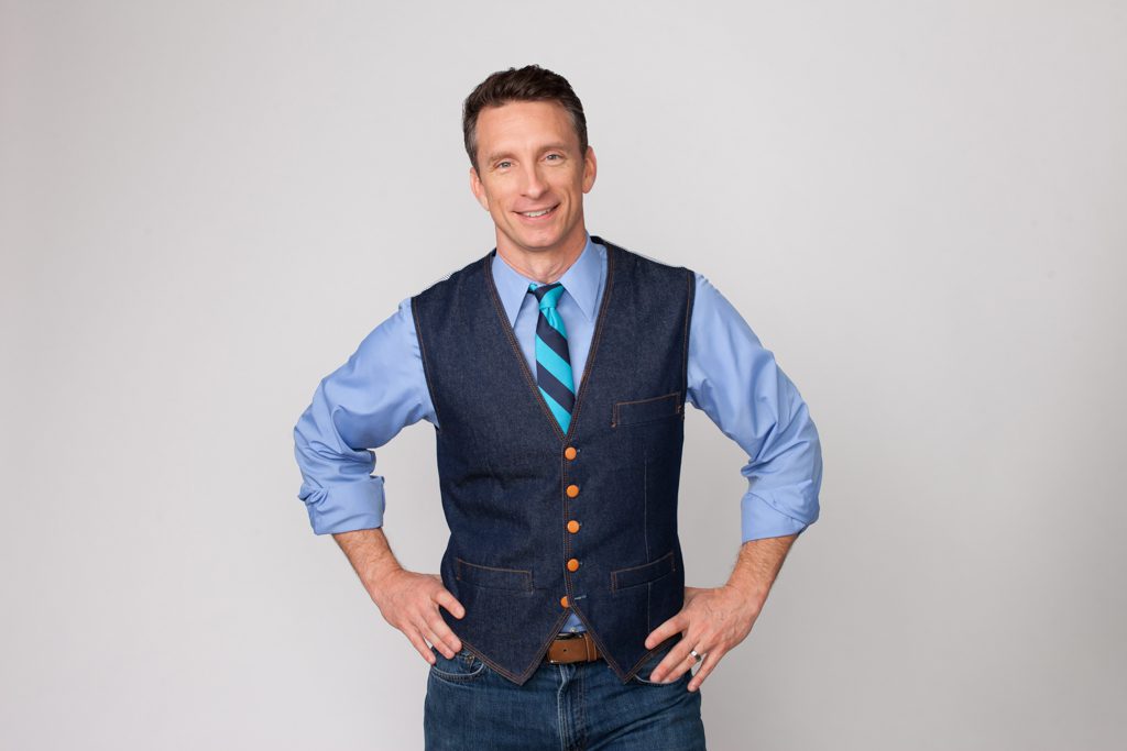 A portrait photo of author Mike Michalowicz dressed in jeans, a denim vest and a blue shirt against a grey background.