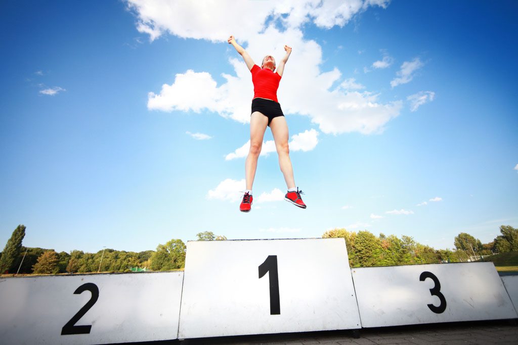 A wide-angle shot of a woman in athletic apparel jumping for joy atop a first-place podium.