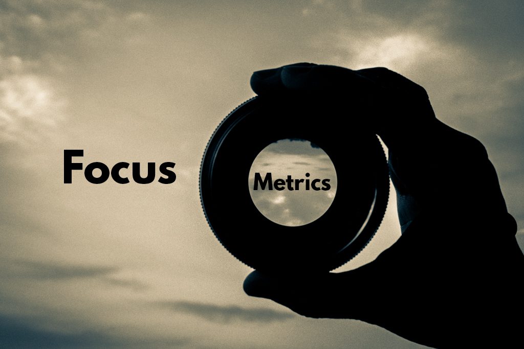 a silhouetted hand holds a camera lens to the sky, with the word "metrics" placed in the center of the lens.