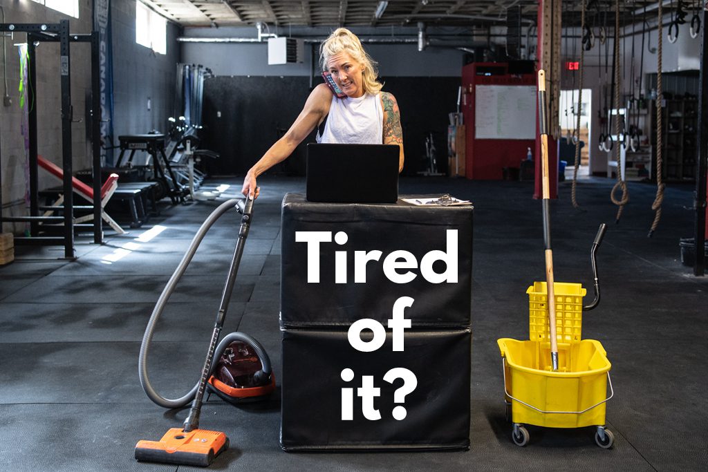 A stressed blond gym owner stands in a gym with a phone at her ear, a vacuum in one hand and a mop bucket nearby.
