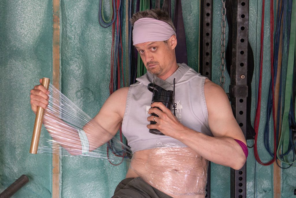 A man dressed in comical fitness attire wraps plastic wrap around his waist.