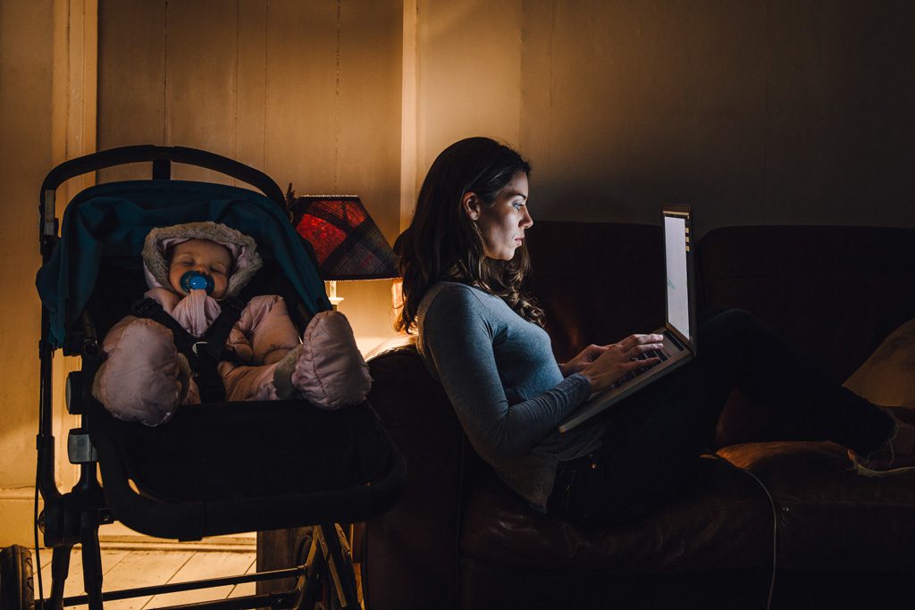 A young mother is working on a laptop on the sofa in her home with her baby sleeping in the push chair next to her.