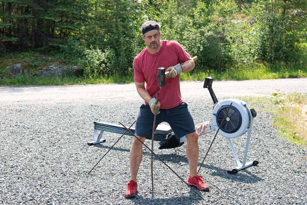 A man in workout gear awkwardly hammers rods in the ground with a sledgehammer.
