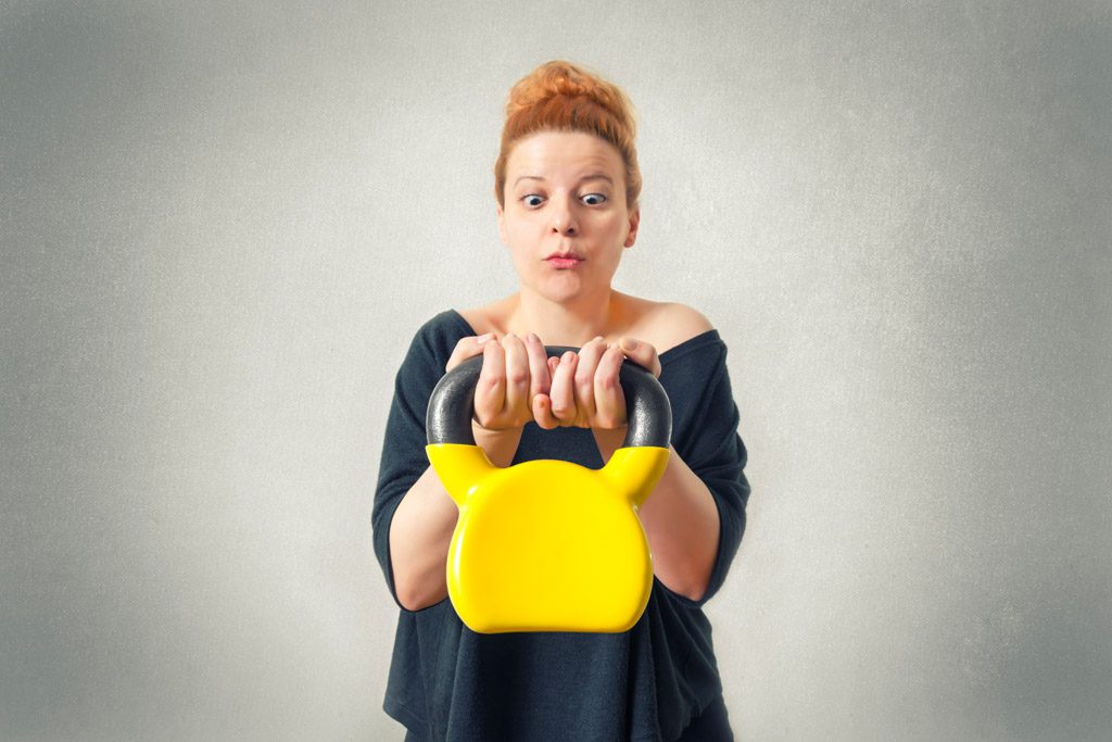A confused woman awkwardly holds a yellow kettlebell in a gym.