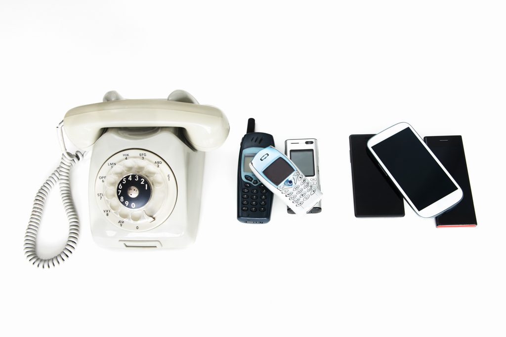 A smartphone, an early cell phone and an old telephone on a white background, from right to left.