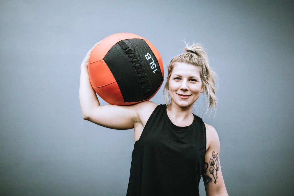 A gym owner with a pony tail smiles as she holds a medicine ball on her shoulder.