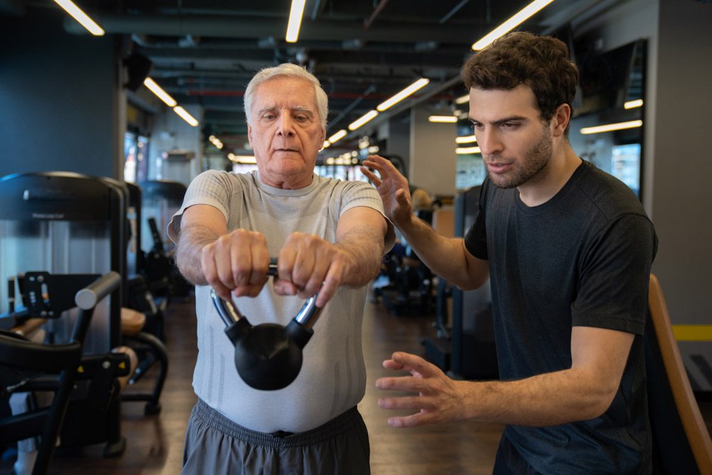 A male personal trainer works with an older client in a commercial gym.