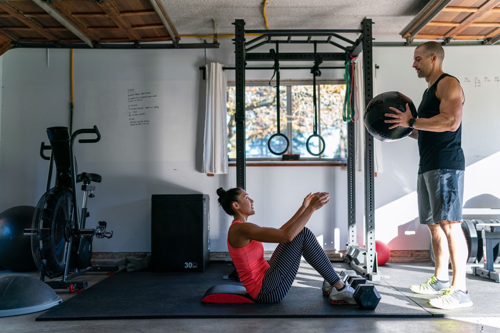 A male personal trainer uses a medicine ball to help a client train her abs in her garage gym.