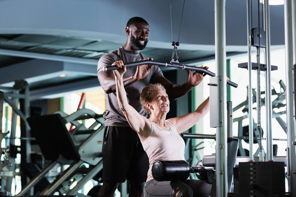 A male personal trainer assists an older female client with a lat pulldown in a gym.