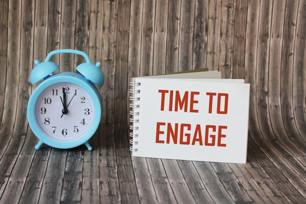 A blue alarm clock sits next to a notepad with "time to engage" written in red letters.