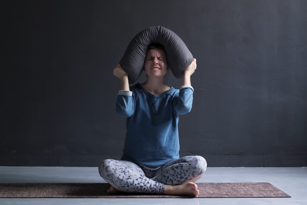 A angry yoga student sits on her mat and scowls as she holds a bolster around her head.