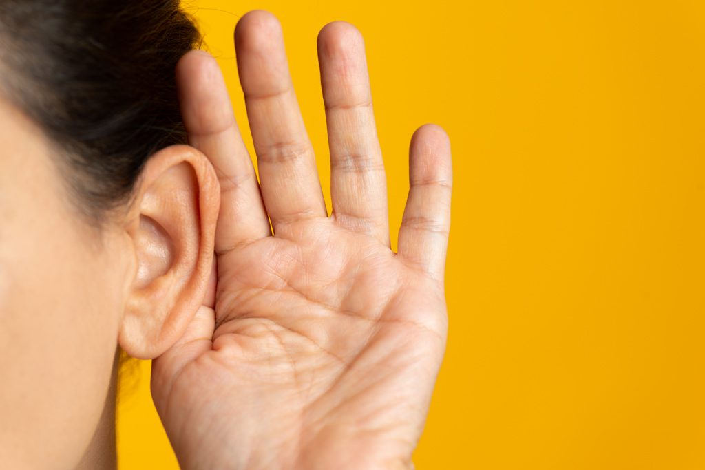 A closeup of a woman cupping her ear to listen on a yellow background.