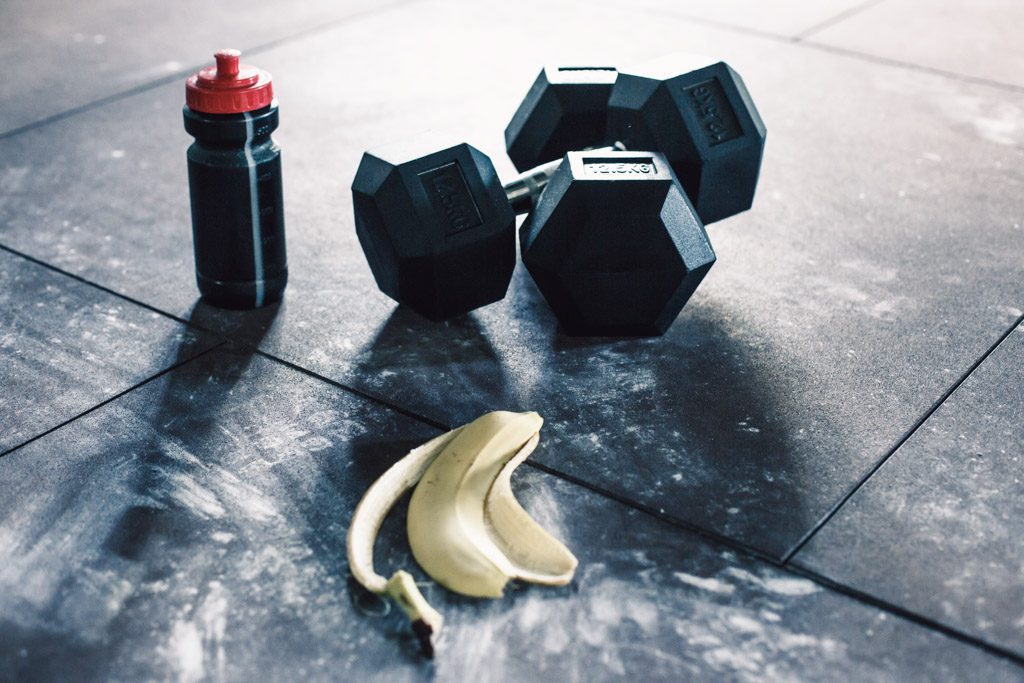 A banana peel sits on a dirty gym floor next to a set of rubber hex dumbbells.