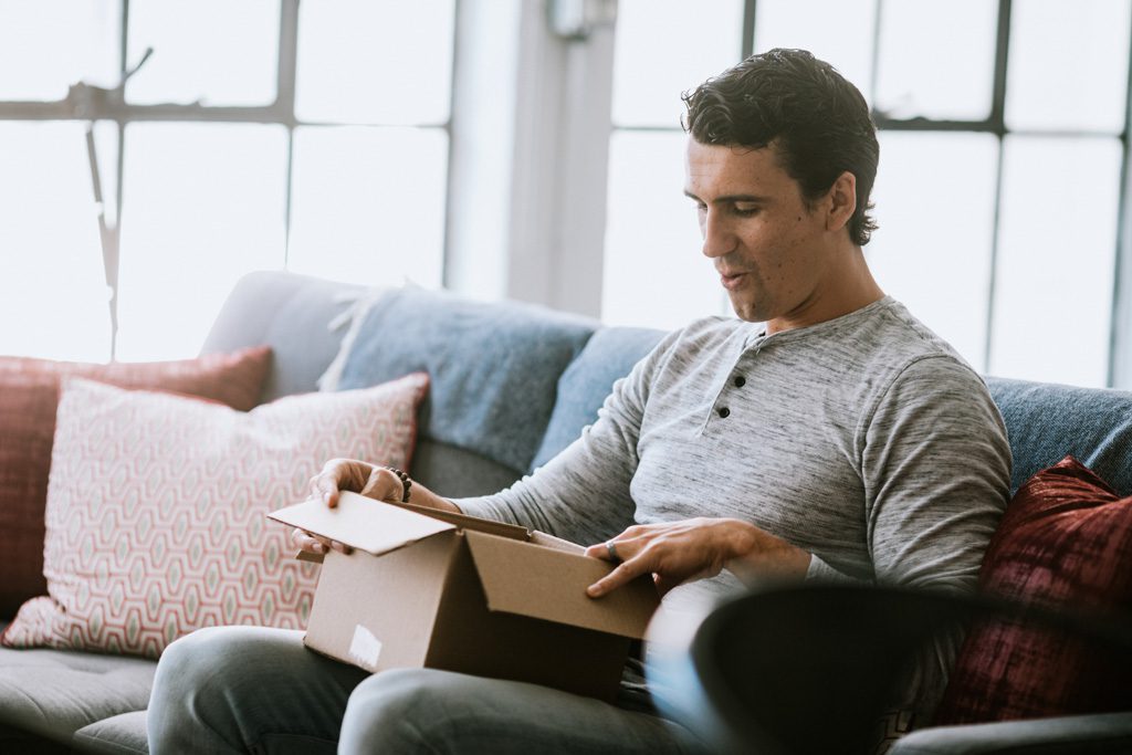 A man seated on a couch looks excited as he opens a package.