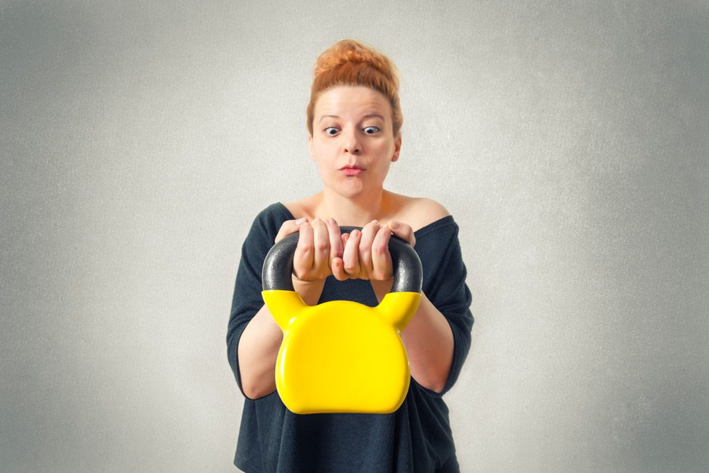 A confused women holds a yellow kettlebell in a gym.