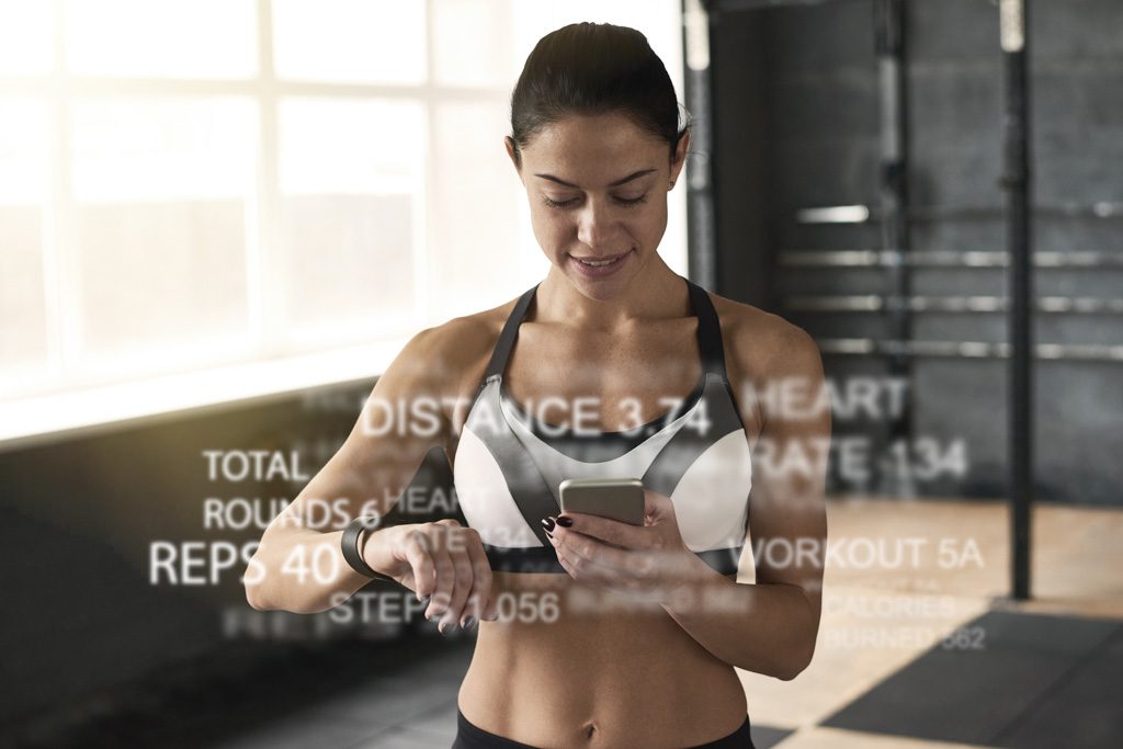 A woman in a sports bra enters workout data on her phone in a gym.