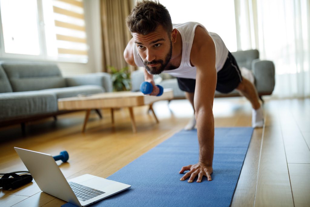 A bearded man does dumbbell rows in a plank as part of an online fitness class.