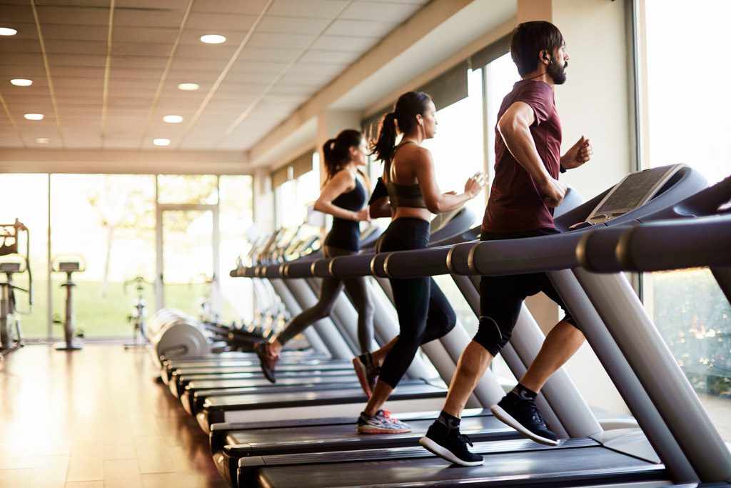 Gym clients run on treadmills facing large windows in a commercial facility.