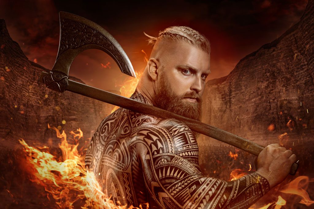 A muscular, tattooed Viking poses with an ax on his shoulder and flames behind him.