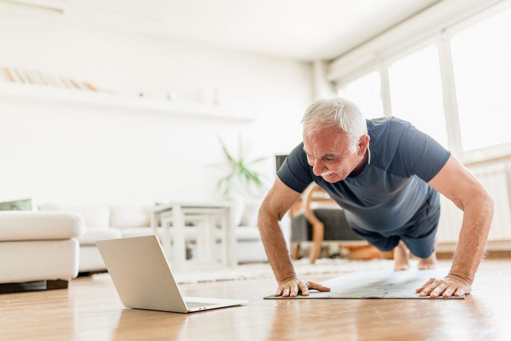 Fitness industry trends: An older gym member performs push-ups in front of a laptop as part of a hybrid program with online coaching.