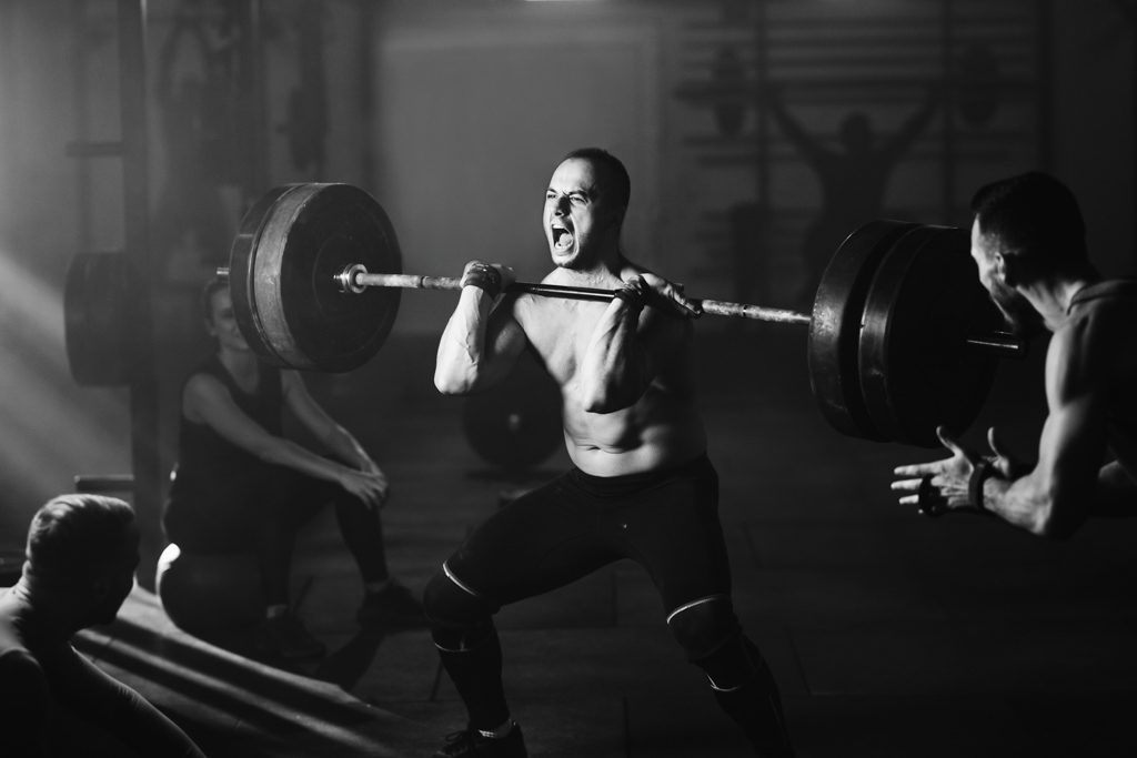 A strong, muscular man yells while performing a power clean in a barbell training facility.