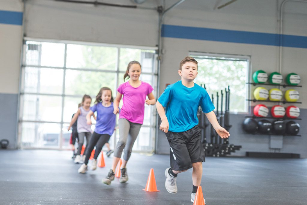 A group of young kids run through a line of orange cones in a gym.