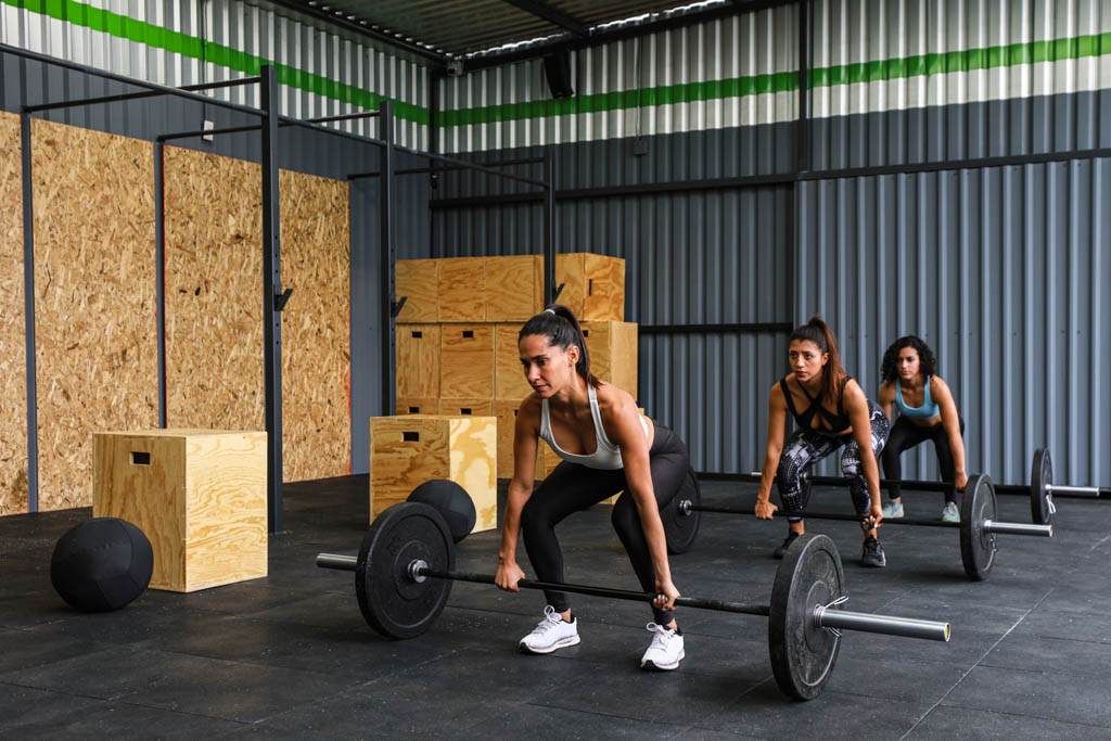 A group of three women prepare to start a competition workout in a gym with deadlifts.