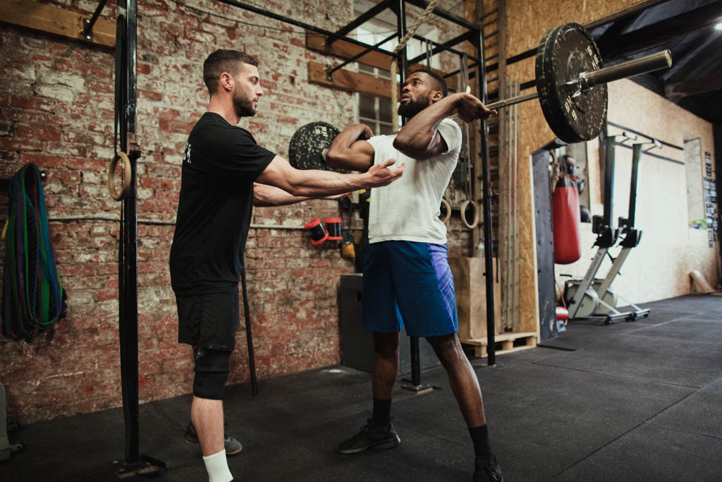 A personal trainer adjusts a client's elbows to help him perform a sound barbell front squat.