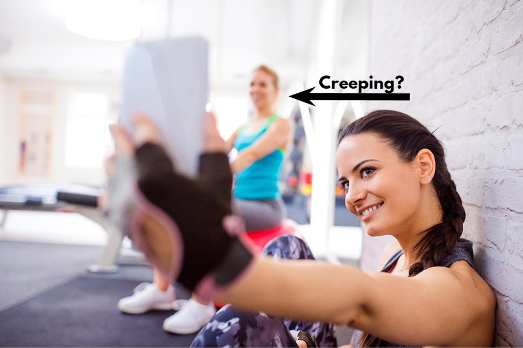 A woman in a gym takes a selfie while another client looks on in the background.